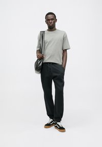 Szara bawełniana koszulka, czarne dresy i czarne sneakersy z białymi akcentami. Model trzyma czarną torbę, ma krótkie rękawy i luźny krój.