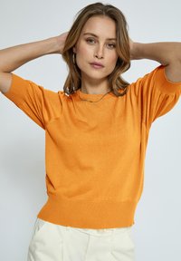 Oranžinė trumpomis rankovėmis megztinis su rausvais kraštais ir apatine dalimi. Minkšta tekstūra, laisvas siluetas ir šiek tiek pūstos rankovės. Dėvima su grietinėlės spalvos kelnėmis.