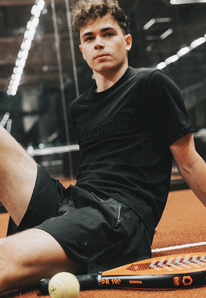 Noor mees istub sisetenniseväljakul mustas spordirõivastuses, oranži padelireketi ja kollase tennispalliga tema jala lähedal.