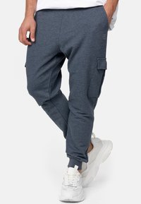 Mężczyzna w ciemnoszarych joggerach cargo i białych sneakersach, z jedną ręką w kieszeni, stojący na tle białego tła.