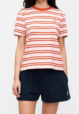 Personne portant un t-shirt à manches courtes rayé horizontalement rose et orange et un short uni bleu marine foncé, les mains détendues sur les côtés.