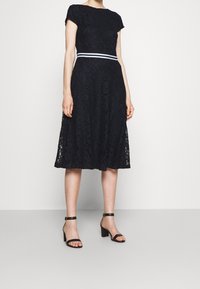 Žena v černých krajkových šatech střední délky s krátkými rukávy, bílým páskem v oblasti pasu a černými sandály na blokovém podpatku s kotníkovým páskem.