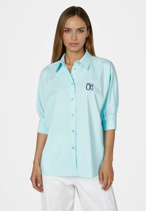 Frau trägt ein hellblaues Hemd mit 3/4-Ärmeln und weiße Hose, steht vor einem schlichten weißen Hintergrund.