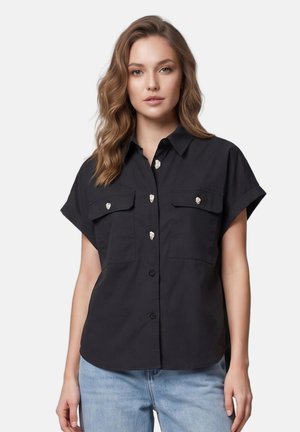 Jeune femme aux cheveux ondulés portant une chemise noire à manches courtes boutonnée avec deux poches sur le devant et un jean bleu clair sur un fond uni.