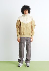 Young person with curly hair stands wearing beige and cream hooded Nike jacket, gray cargo pants, and gray and white sneakers on green floor.