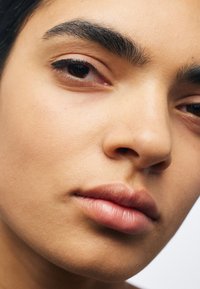 Gros plan sur un visage à la peau marron clair, avec des sourcils bien définis, des lèvres douces et un maquillage des yeux subtil. Texture lisse et teint naturel.