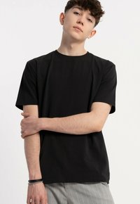 Młody mężczyzna z krótkimi, kręconymi włosami, ubrany w czarną koszulkę i szare w paski spodnie, stoi z jedną ręką skrzyżowaną na drugiej.