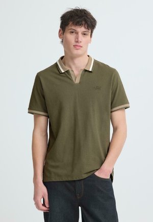 Jeune homme debout, une main dans la poche, portant un polo vert olive avec des bordures beiges et un jean foncé, sur un fond uni.