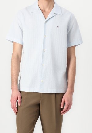 Homme portant une chemise bleu clair à manches courtes avec un petit logo sur la poitrine et un pantalon marron, debout devant un fond uni.