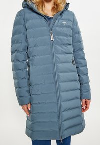 Pitkä, pörröinen talvitakki pehmeästä, tekstuurillisesta sinisestä kankaasta, jossa on huppu, sivutaskut ja edessä vetoketjusuljenta. Rinnassa on logo.