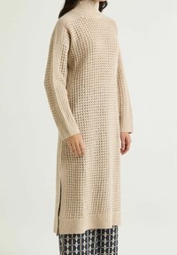 Vestito maglione beige con collo alto, texture a coste e spacchi laterali. Caratterizzato da una vestibilità ampia e maniche lunghe.