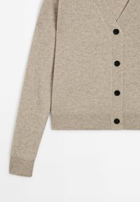 Cardigan en tricot beige avec un col en V, boutons noirs, poignets côtelés et une taille raccourcie. Texture douce avec un léger motif chiné.