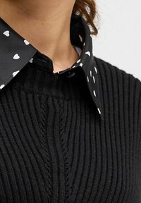 Maglione nero a coste con un colletto nero caratteristico decorato con un motivo a cuori bianchi. Il colletto presenta una chiusura a bottone e uno strato interno.