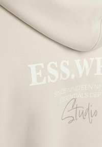 Šviesiai smėlinis hoodie, pagamintas iš minkštos medžiagos; turi baltą spausdintą tekstą ir ranka rašytą detalę, pabrėžiančią prekės ženklą ir būtinų daiktų skyrių.