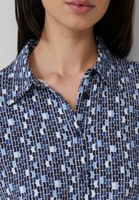 Blau gemusterte Knopfleiste mit Kragen, mit abwechselnd marineblauen und hellblauen abstrakten Formen und zwei sichtbaren dunklen Knöpfen.