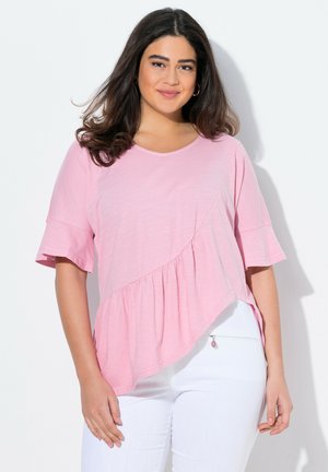 Roze top met korte mouwen en een gebogen zoom, voorzien van een diagonale paneelontwerp en een geplooid detail. In combinatie met witte broeken.