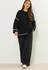 Maglione nero lavorato a maglia con cuciture decorative bianche, vestibilità morbida, scollatura tonda, abbinato a pantaloni neri a gamba larga e sneakers bianche.