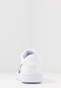 Vit Reebok-sneaker sedd bakifrån, med marinblå och röda sidodetaljer och en texturerad vit sula på en vit bakgrund.