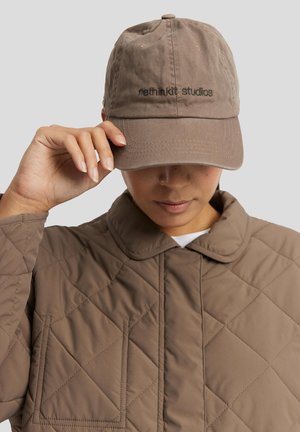 Osoba koja nosi smeđu prošivenu jaknu i smeđu kapu s vezom "nethin'kit studios", prilagođava vizir kape desnom rukom.