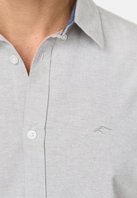 Camisa abotonada de color gris claro con un tejido texturizado, cuello interior azul en contraste y un sutil logo bordado en el pecho.