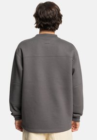 Sudadera gris con un cuello redondo y mangas largas, que presenta una costura horizontal en la parte posterior y puños y dobladillo acanalados.