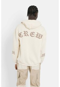 Felpa leggera beige con "CREW" in grassetto marrone sul retro, grande cappuccio e polsini a coste. Indossata con pantaloni cargo beige.