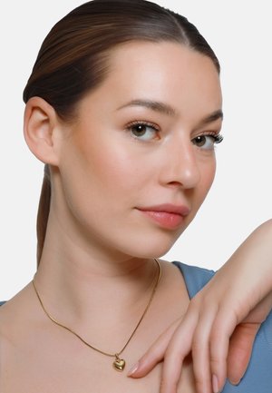 Junge Frau mit leichtem Make-up, die eine goldene Herz-Anhänger-Halskette und ein blaues Oberteil trägt, posiert mit ihrer Hand nahe am Gesicht.