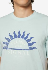 Šviesiai mėlyna medvilninė marškinėlių su retro saulės grafika tamsiai mėlyna spalva, turinčia bangomis panašius spindulius, ir užrašu "Outdoor Club" bei "Signal EST 1967" po ja.