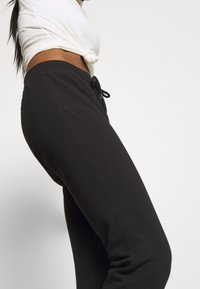 Kvinna klädd i svarta dragsko-byxor och en vit kortärmad topp med knyt framtill, mot en enfärgad ljus bakgrund.