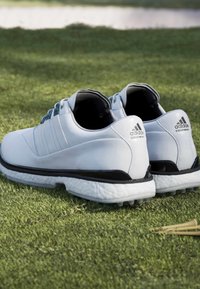 Bílé syntetické golfové boty s černými akcenty, šněrovací design, texturovaná podrážka a značka Adidas na patě. Vhodné na travnaté povrchy.