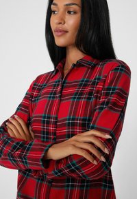 Camisa de franela roja con patrón a cuadros en negro y verde, cuello abotonado, mangas largas y textura suave. Brazos cruzados sobre el pecho.