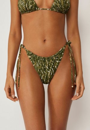 Kvinde iført grøn tie-dye bikini med bånd til at binde i siden på trusserne og trekants-top, stående mod en ensfarvet lys baggrund.