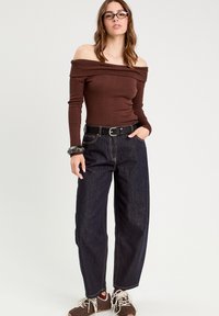 Brun ribbet off-shoulder top parret med højtsiddende mørkeblå jeans. Tilbehør inkluderer et sort bælte og et tekstureret armbånd.