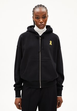 Woman with braided hair wearing a black zip-up hoodie and white turtleneck, standing against a plain white background.