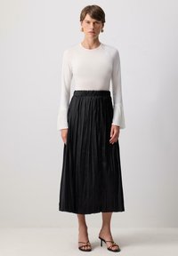 Vit långärmad topp med veckade ärmslut, kombinerad med en svart, veckad kjol med ett samlat midjeband och svarta klacksandaler.