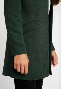 Cardigan long texturé vert avec une coupe légèrement ample, doté d'une poche sur le côté gauche et d'un ourlet droit.