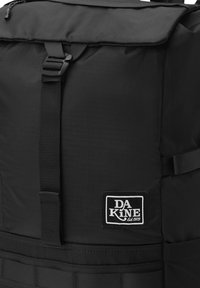 Svart ryggsäck med en strukturerad tyg, justerbara remmar, en övre lucka och en logotyp på lappen med "DA KINE Est. 1979." Horisontella webbing-accenter.