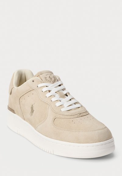 Sneaker en suède beige avec des lacets blancs, à bout rond et détails du logo sur le côté. Semelle en caoutchouc blanc épais avec des accents texturés subtils.