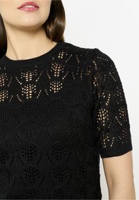 Juodas trumpas rankovėmis megztinis su atviru raštu, kuris turi gėlių ir geometrinių raštų, su banguotu apykakle ir minkšta tekstūra.