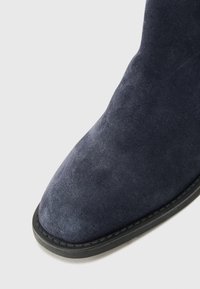 Bottine en daim bleu marine avec une texture lisse, bout arrondi et semelle en caoutchouc noir. Couture détaillée le long du bord de la semelle.