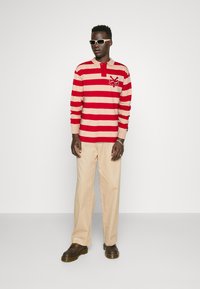 Red and beige striped long-sleeve polo shirt with a collar and emblem, paired with beige trousers and brown shoes. White sunglasses worn.