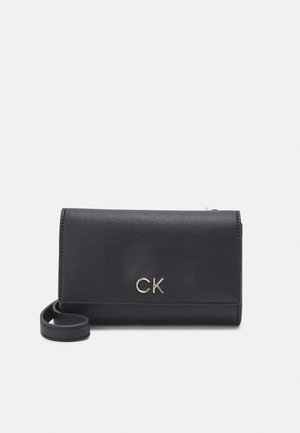 Zwarte leren polstasje met goudkleurig "CK"-logo aan de voorkant en een korte riem aan de zijkant, geplaatst tegen een lichte achtergrond.