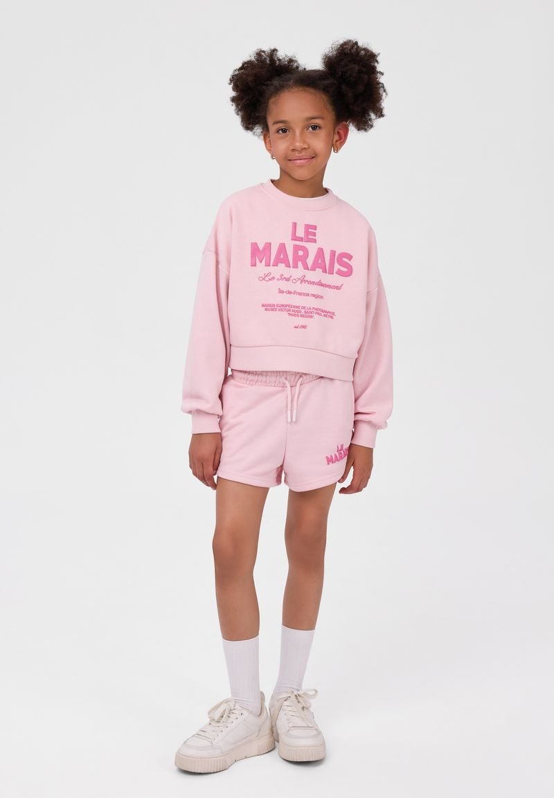 Fille avec des touffes afro portant un sweat-shirt rose "Le Marais" et un short assorti, des chaussettes blanches et des baskets blanches, debout devant un fond uni.