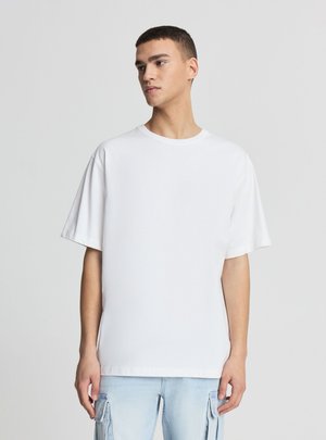 Giovane uomo con capelli corti che indossa una T-shirt bianca oversize semplice e jeans cargo azzurri chiari, guarda a sinistra su uno sfondo neutro.