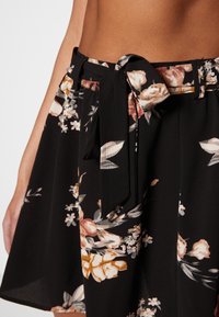 Jupe noire à fleurs avec une ceinture à nouer, présentant un design en couches mettant en avant des fleurs roses et oranges sur un tissu doux et fluide.