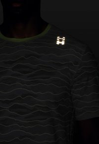 Musta ja vihreä lyhythihainen t-paita, jossa on viimeistelty aaltojen kuvio ja heijastava logo olkapäällä, lähikuvassa.