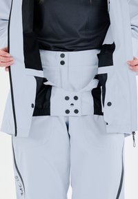 Ljusblå skidjacka med svart inre foder, med tryckknappar och ventilation. I kombination med matchande skidbyxor, accentuerade med logotyper.