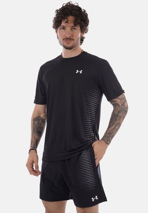 Uomo con tatuaggi che indossa abbigliamento sportivo nero Under Armour con motivo a pois, in piedi davanti a uno sfondo bianco uniforme.