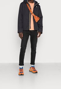 Tmavě námořnická bunda s kapucí, oranžová taška přes rameno, světle oranžové tričko, černé džíny a vícebarevné tenisky s modrými a oranžovými detaily.