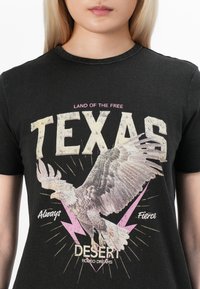 Černé triko s velkým bílým nápisem "TEXAS", grafikou létajícího orla, růžovými blesky a frázemi "Krajina svobody", "Vždy odvážní" a "Sny z pouštního rodea."